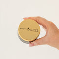 Hand holding a gold container with 'Archie Lloyd's Hair Extensions' branding on a light beige background